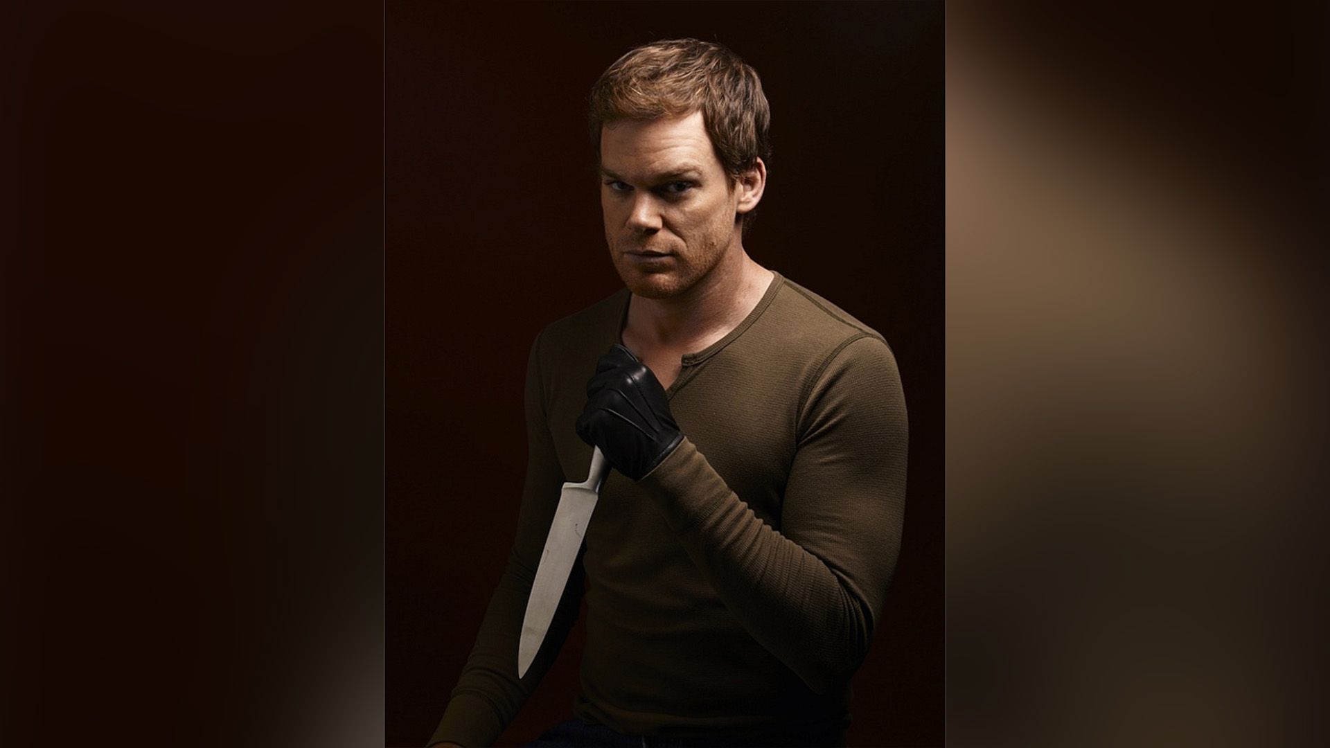 Michael C. Hall con una giacca marrone e guanti neri che tiene un coltello in mano