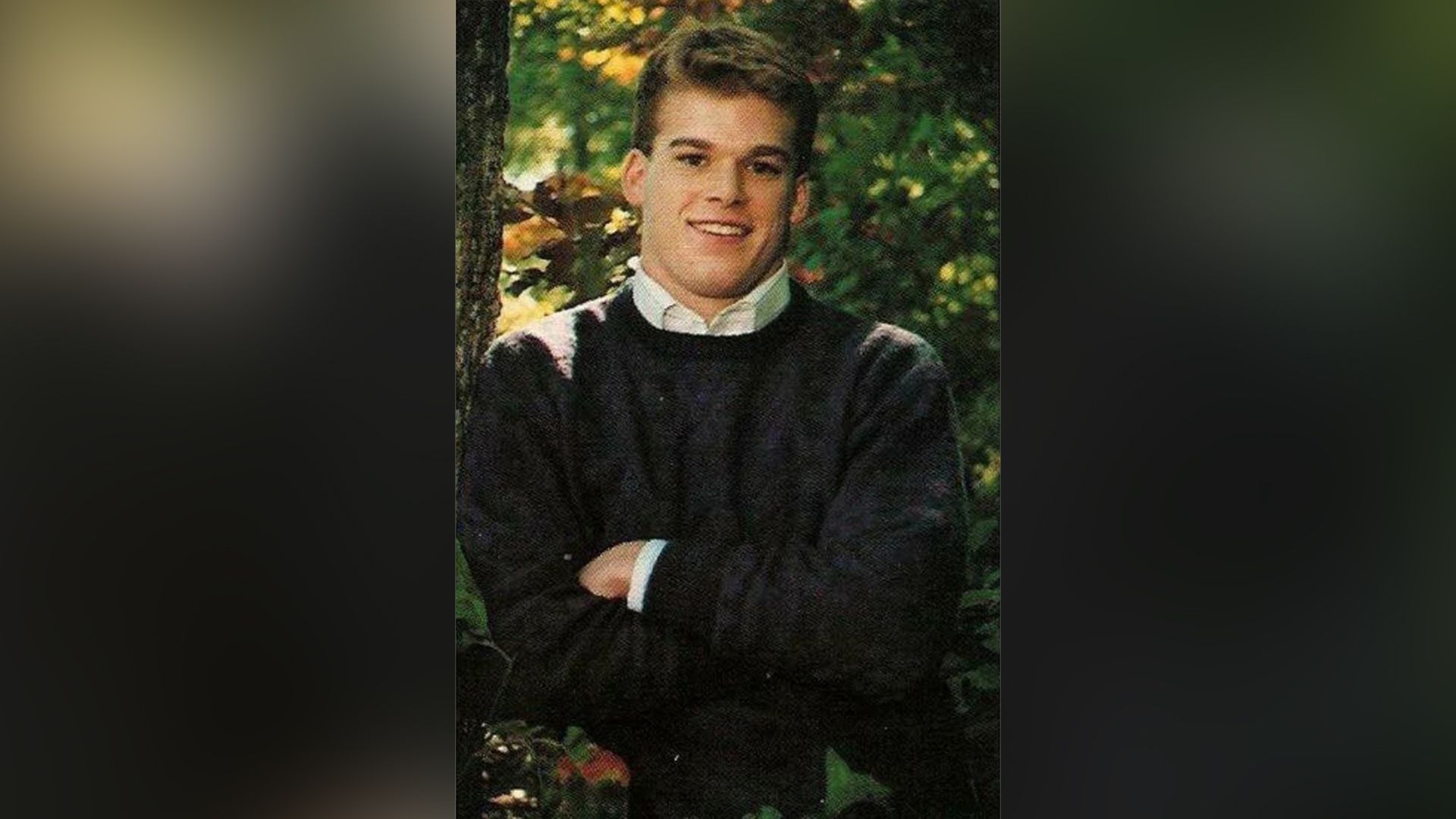 Il giovane Michael C. Hall sta in piedi con le braccia incrociate sul petto, sorridendo ampiamente