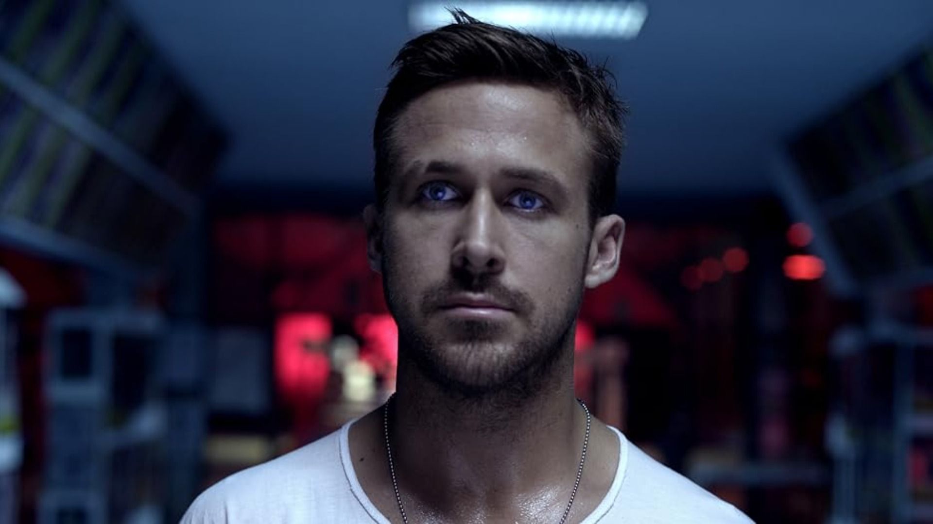 L'attore Ryan Gosling in maglietta bianca in piedi contro uno sfondo sfocato