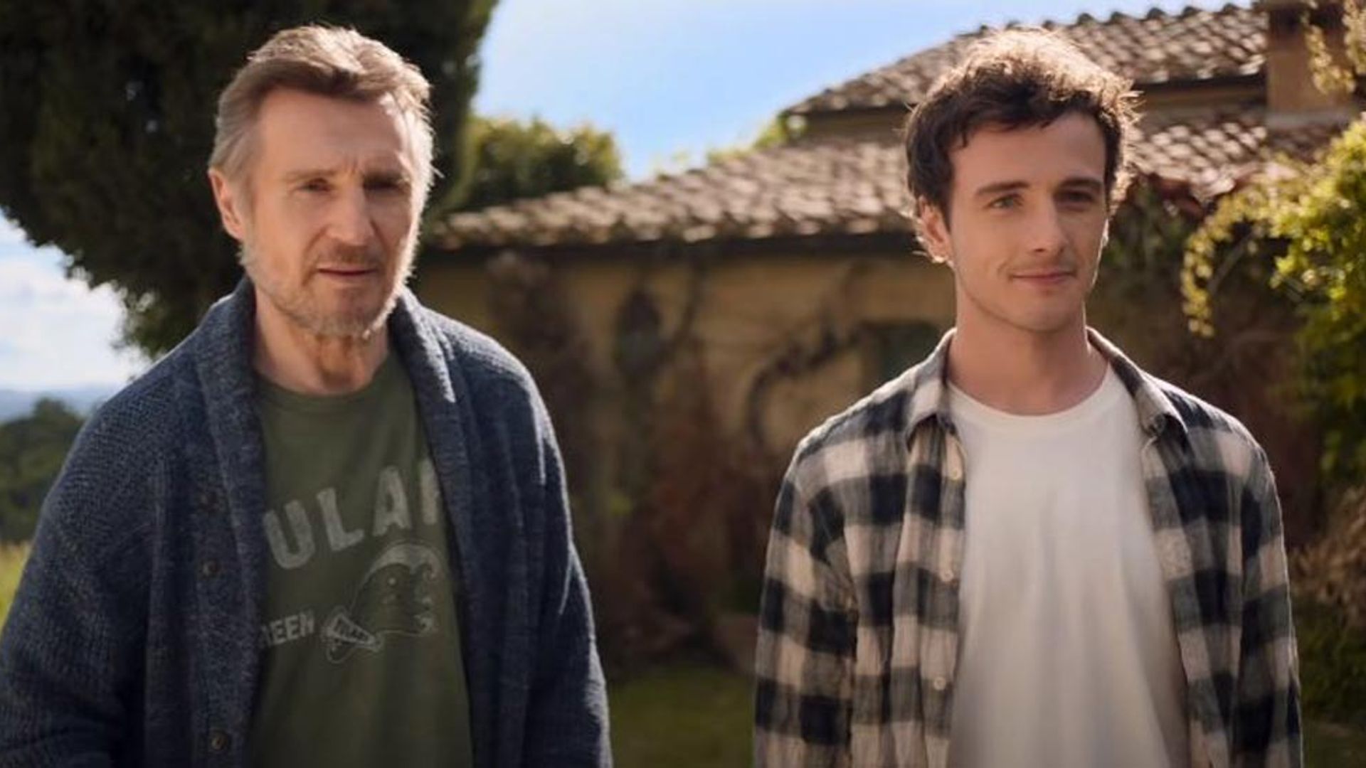 Liam Neeson e Michael Richardson stanno fianco a fianco davanti a una casa con tetto di tegole in una giornata di sole