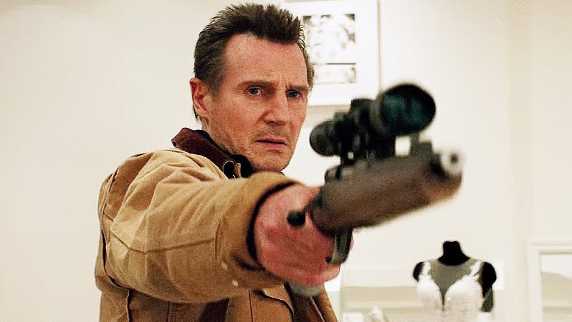Liam Neeson in una giacca beige punta un fucile con mirino, con un manichino in abito da sposa dietro di lui