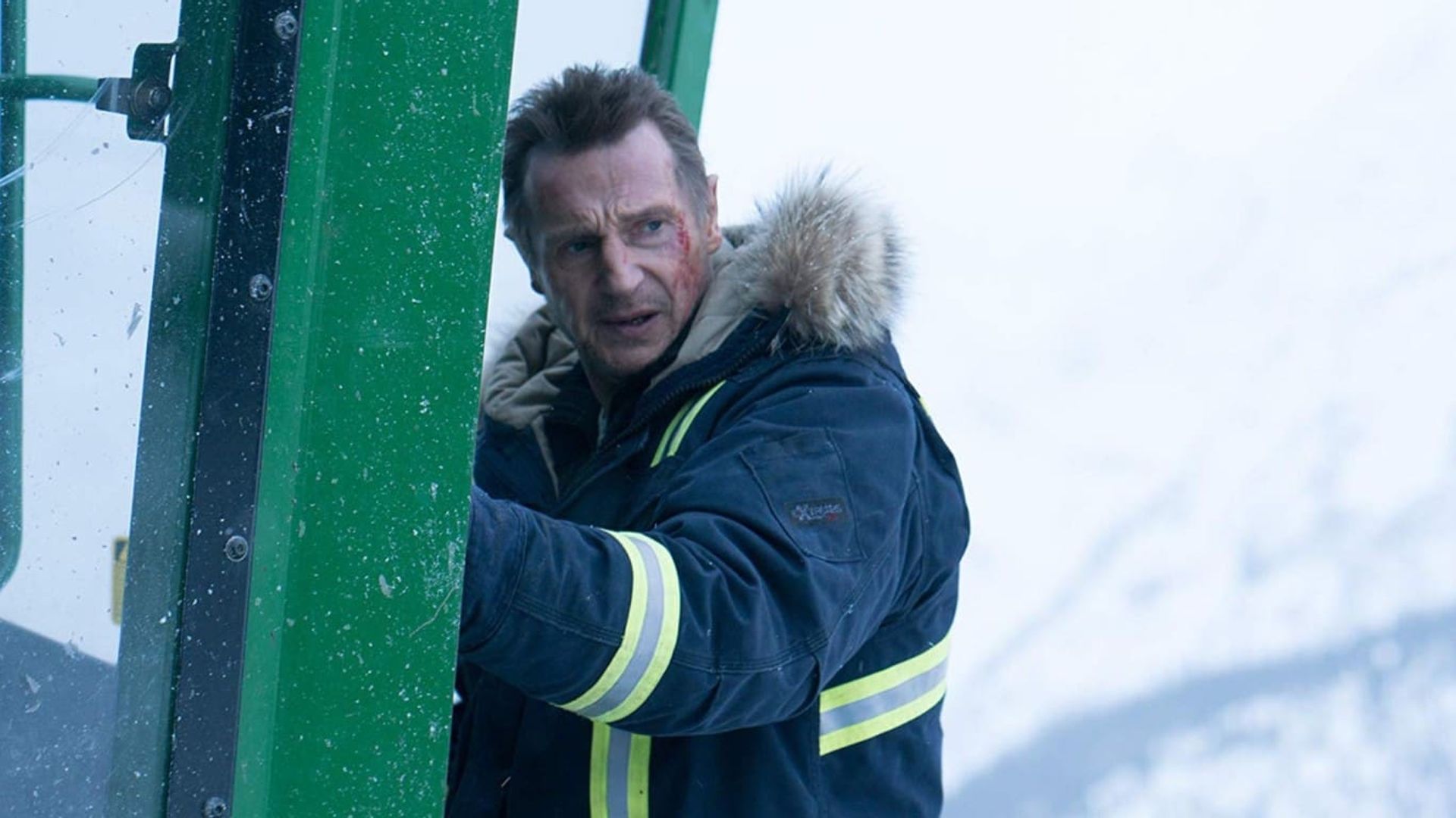 Liam Neeson in una giacca invernale con cappuccio afferra la porta di una baita verde con un paesaggio innevato sullo sfondo
