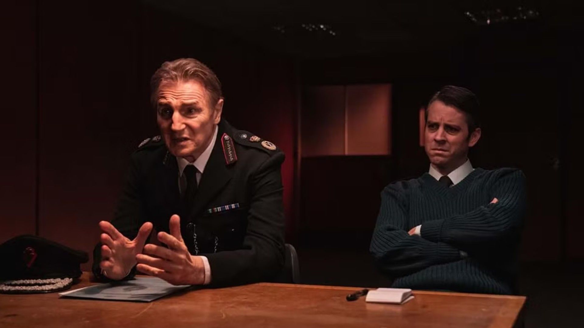 Liam Neeson in uniforme da poliziotto seduto a un tavolo di interrogatorio, scena dalla serie "Derry Girls"