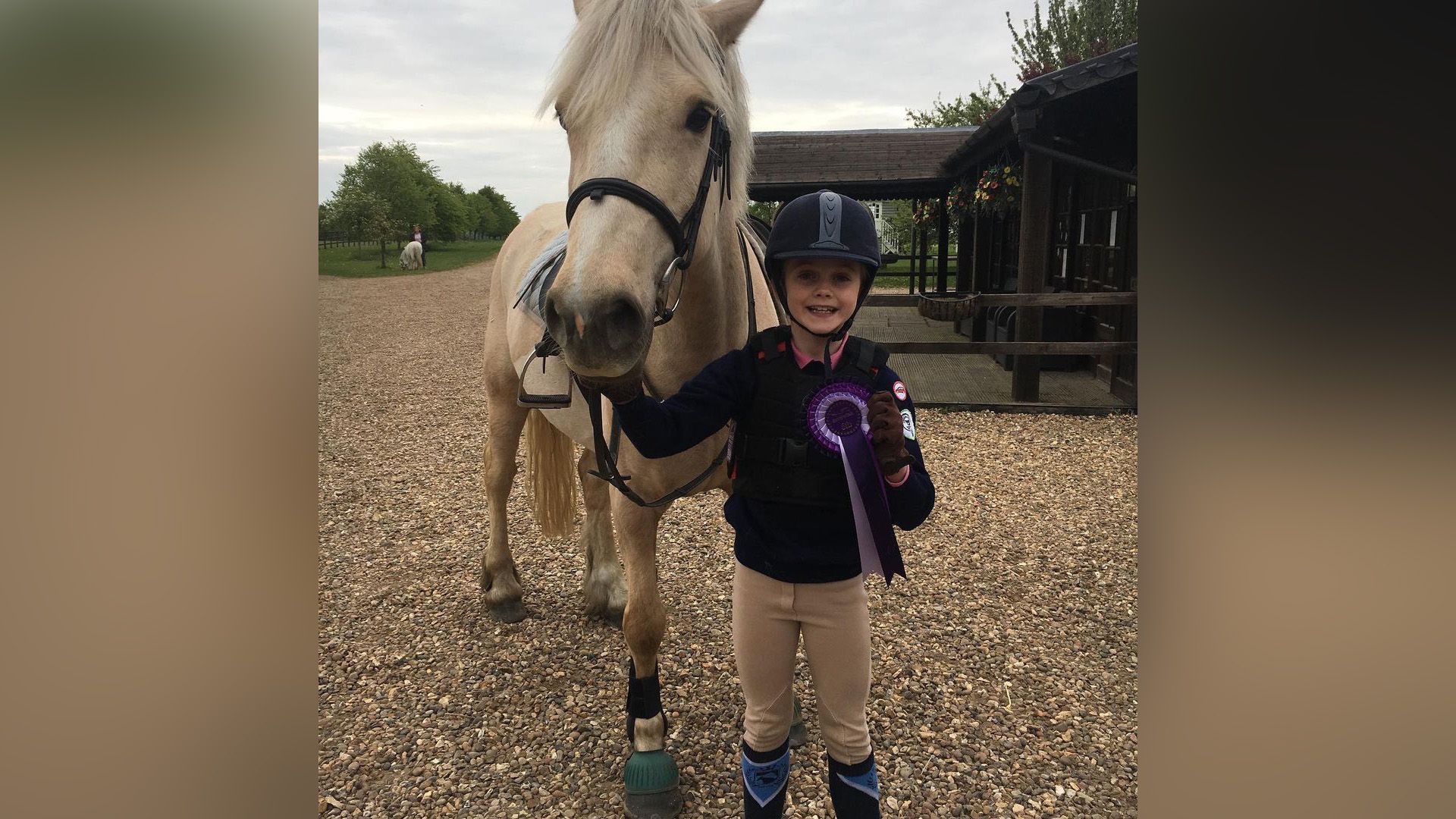 Evie Templeton da bambina, vestita con abbigliamento da equitazione e casco, in piedi accanto a un cavallo mentre tiene in mano un nastro premio viola