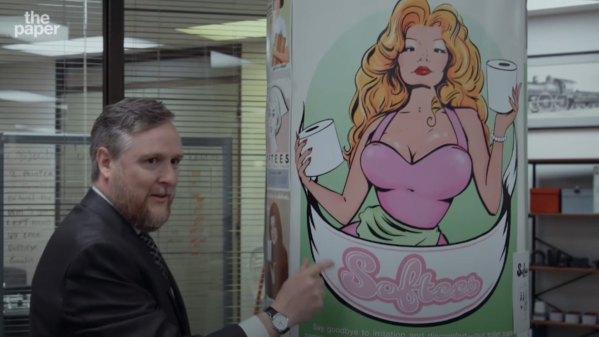 Un uomo in giacca e cravatta accanto a un poster con una donna bionda disegnata che tiene rotoli di carta igienica