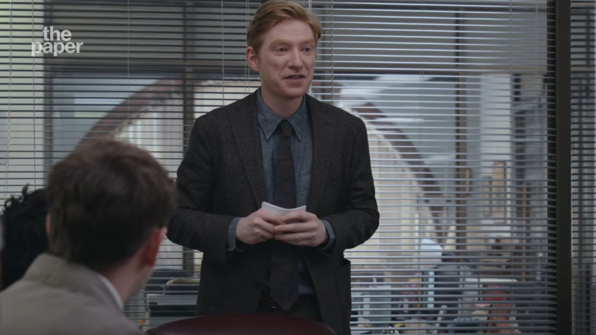 Domhnall Gleeson in giacca e cravatta in piedi in una sala conferenze