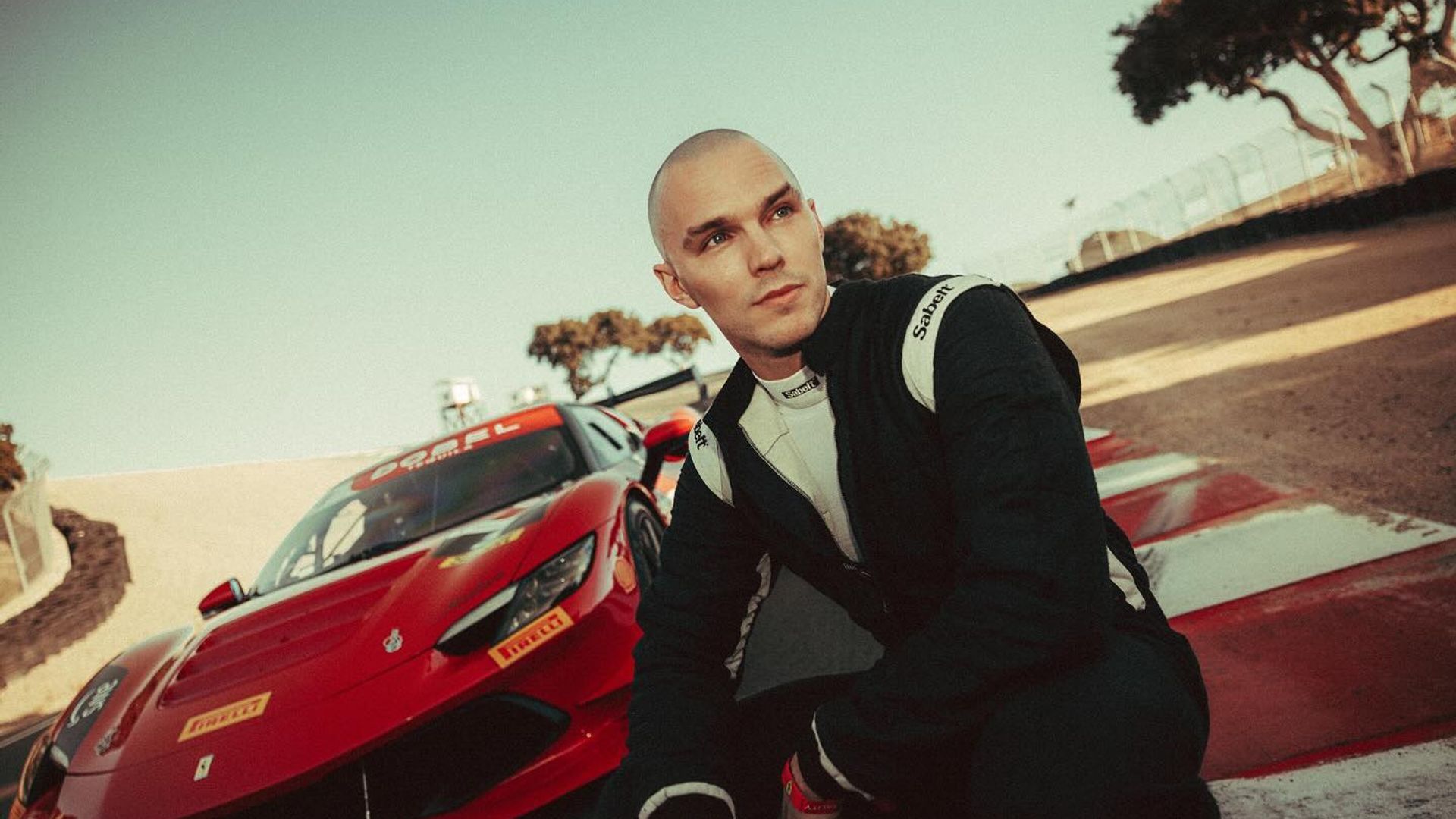 Nicholas Hoult in tuta da corsa seduto sulla pista con una Ferrari rossa sportiva dietro di lui