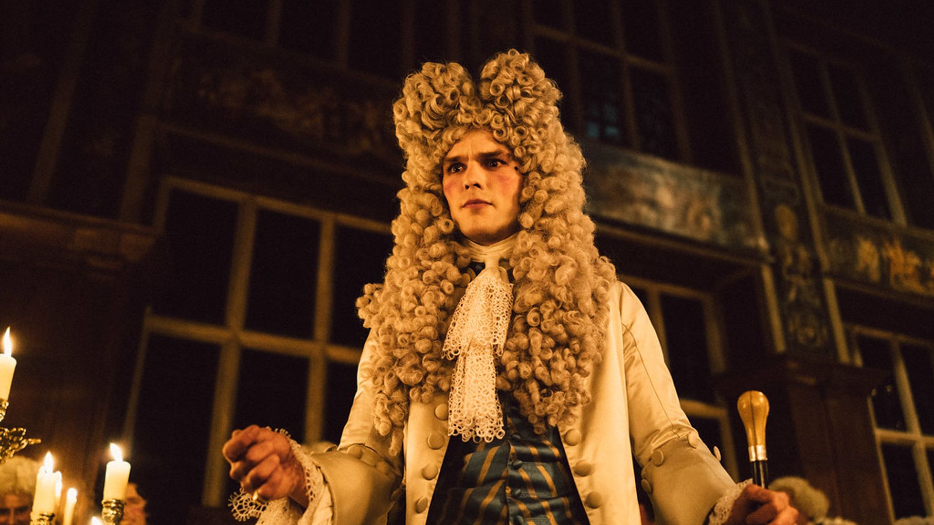 Nicholas Hoult con una parrucca e un costume d'epoca del XVIII secolo con un bastone da passeggio in piedi in una sala illuminata da candele