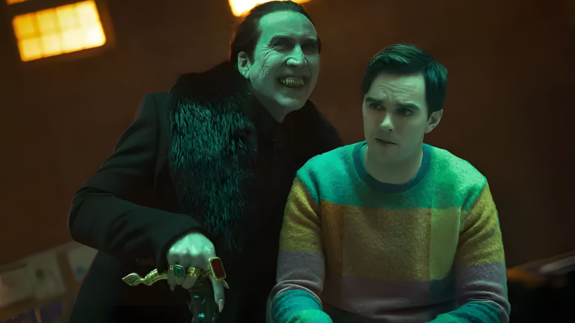Nicolas Cage in abiti neri con anelli incombe minacciosamente su Nicholas Hoult in un maglione colorato