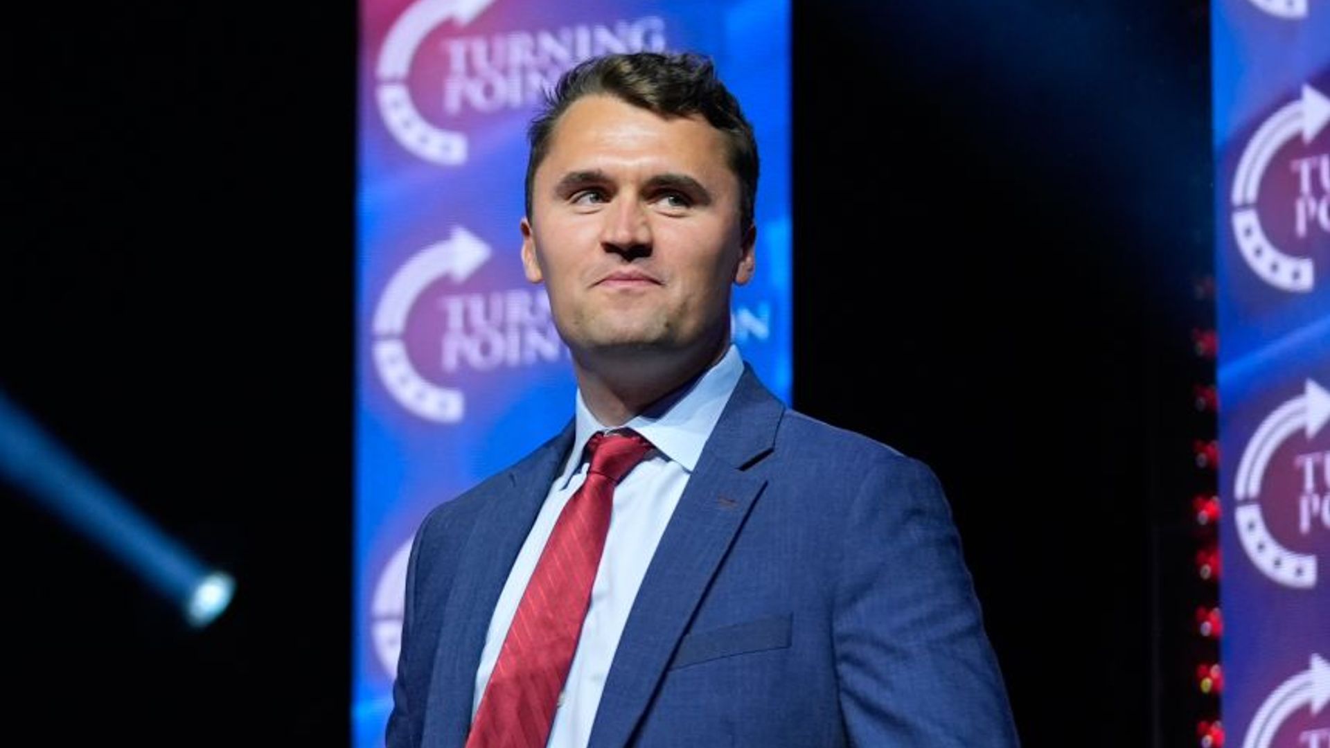 Charlie Kirk in posa fiera su sfondo scuro indossando un completo blu