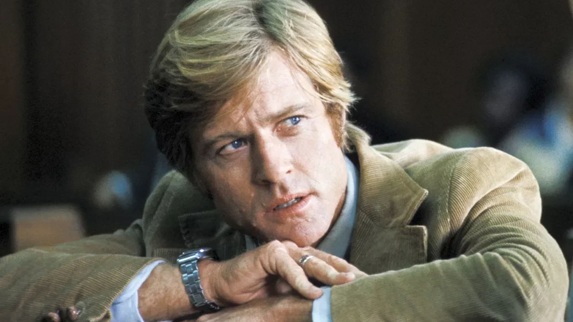 Robert Redford in giacca marrone con un orologio al polso, appoggiato sulle mani mentre guarda di lato