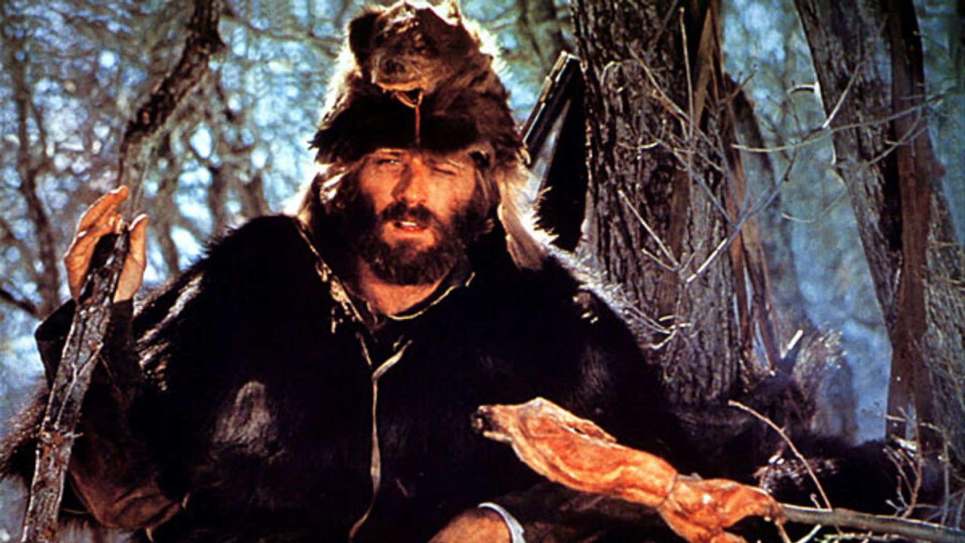 Robert Redford in abiti di pelliccia e un berretto di testa di bestia che griglia carne su un fuoco da campo nella foresta, scena dal film "Jeremiah Johnson"