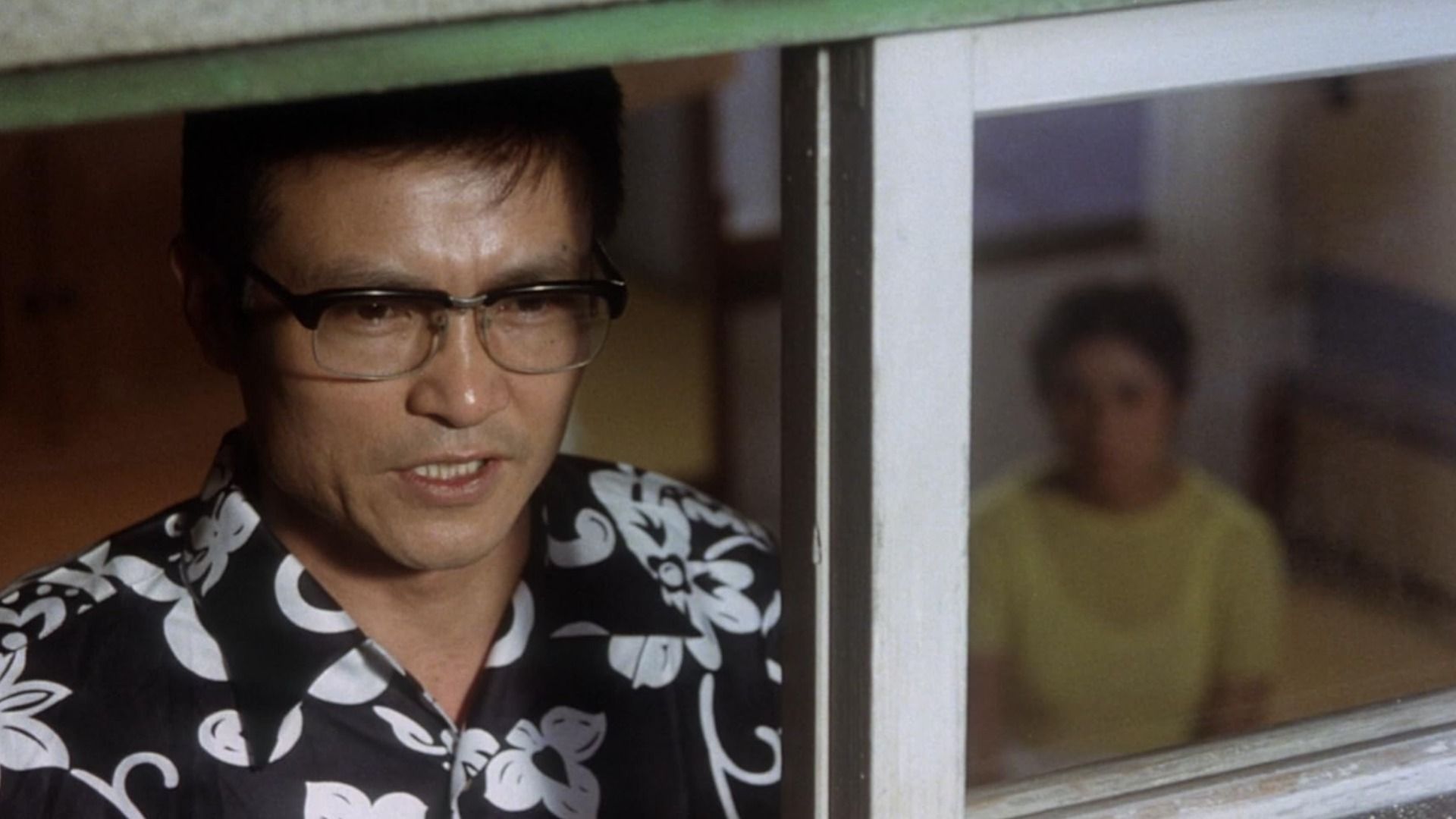 Un uomo con gli occhiali e una camicia nera sta davanti a una finestra con una ragazza seduta dietro di lui, sfocata