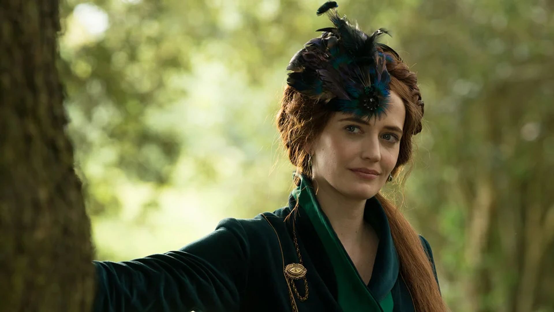 Eva Green in un abito verde scuro con ornamenti di piume sulla testa sta in piedi accanto a un albero su uno sfondo di foresta