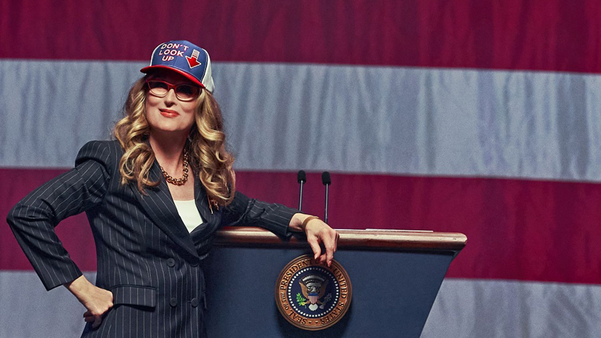 Meryl Streep in abito e berretto con bandiera americana in piedi contro uno sfondo della bandiera statunitense, appoggiata a un podio