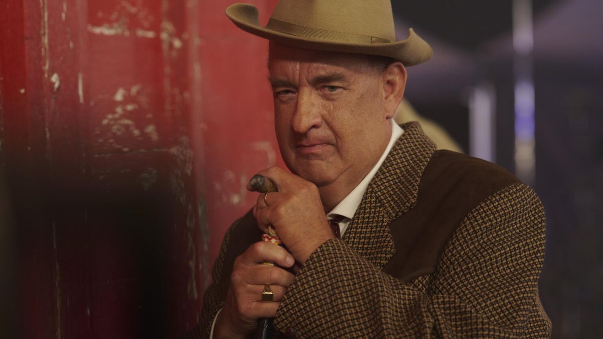 Tom Hanks in abito marrone e cappello si appoggia a un bastone, guardando seriamente verso la telecamera