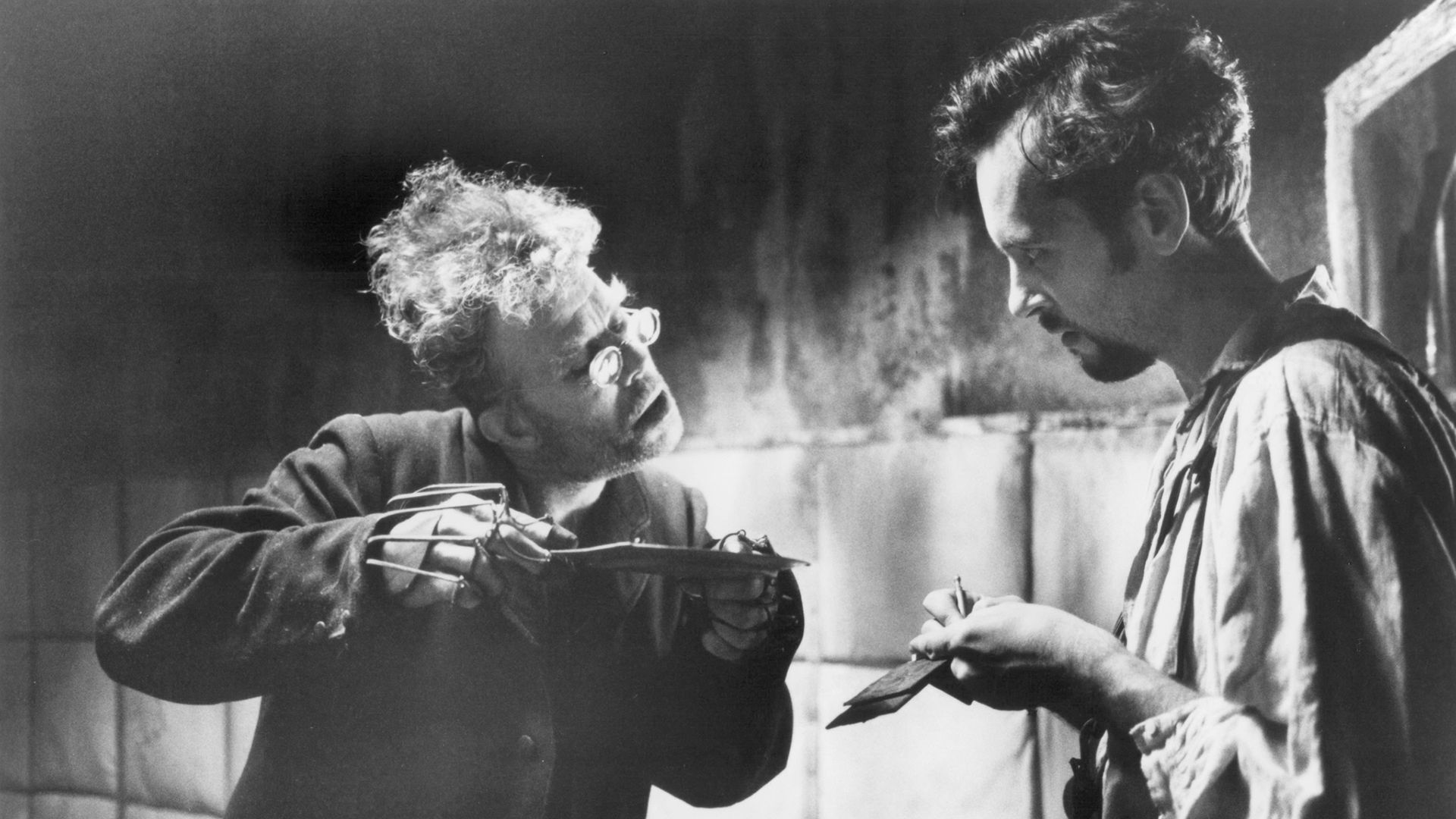 Tom Waits con occhiali rotondi e protesi alle dita tiene un piattino tra le mani, accanto a lui c'è un giovane uomo con un bloc notes