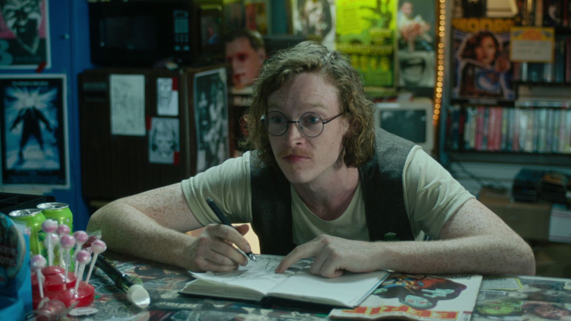 Caleb Landry Jones con occhiali e gilet seduto a un tavolo con un blocco note, disegna e guarda di lato