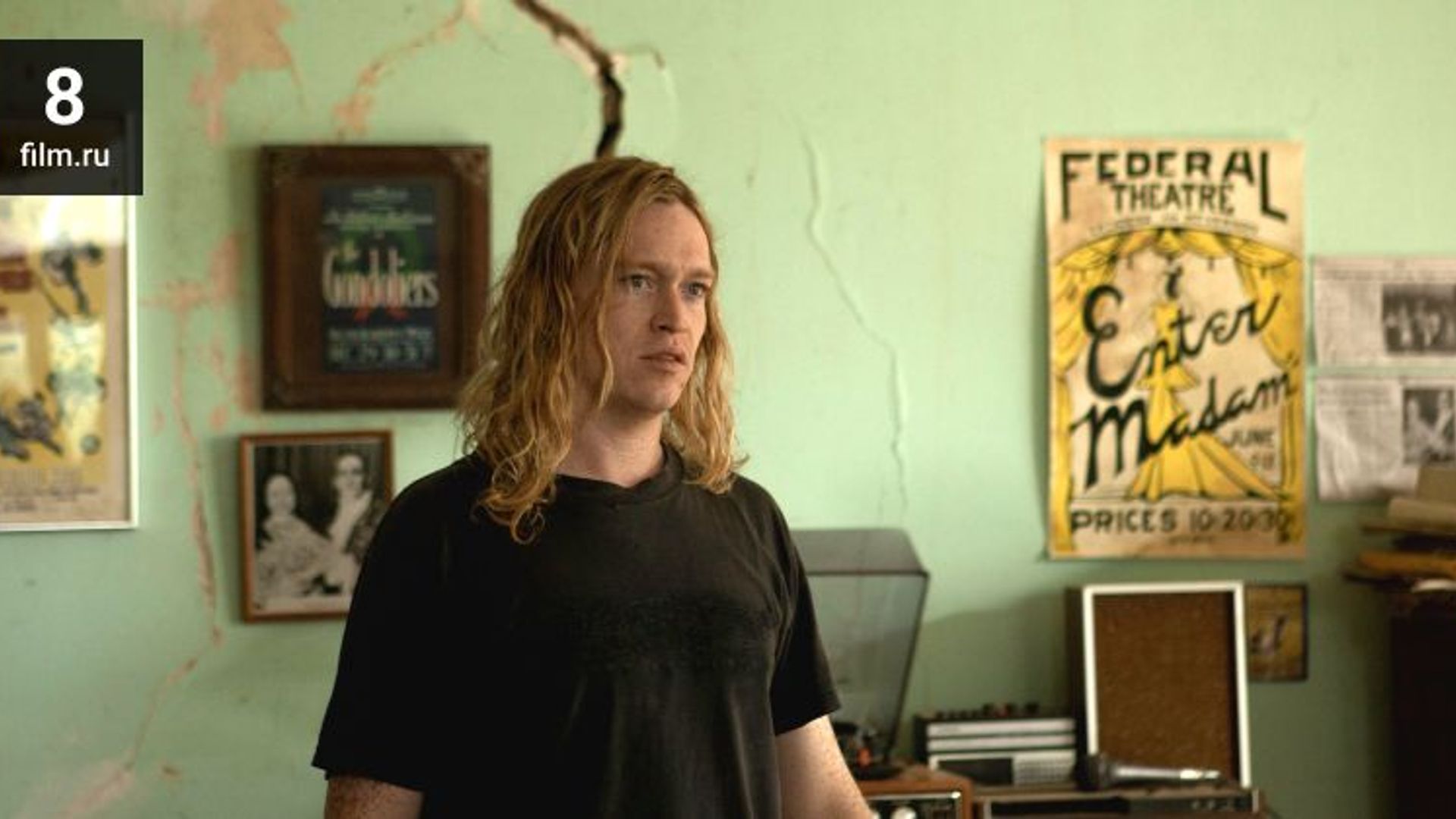 Caleb Landry Jones con lunghi capelli biondi in una maglietta nera si trova in una stanza con poster alle pareti