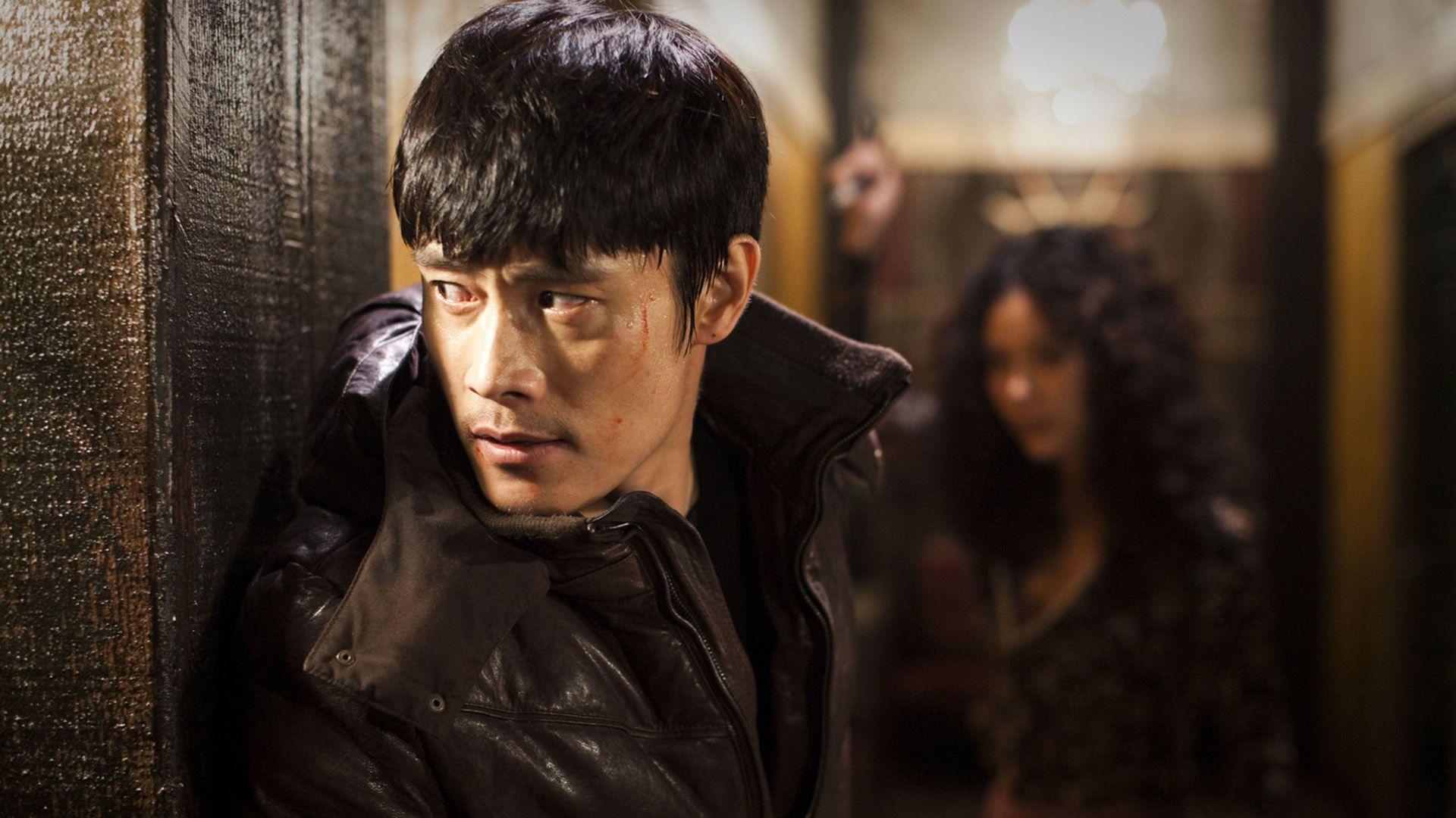 Lee Byung-hun in giacca nera sbircia dall'angolo di un edificio
