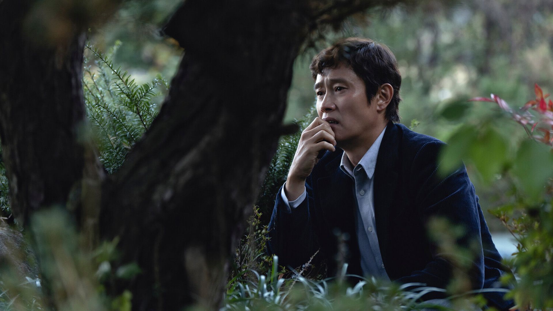 Lee Byung-hun in blazer blu seduto sull'erba tra gli alberi, con un'espressione disperata sul volto