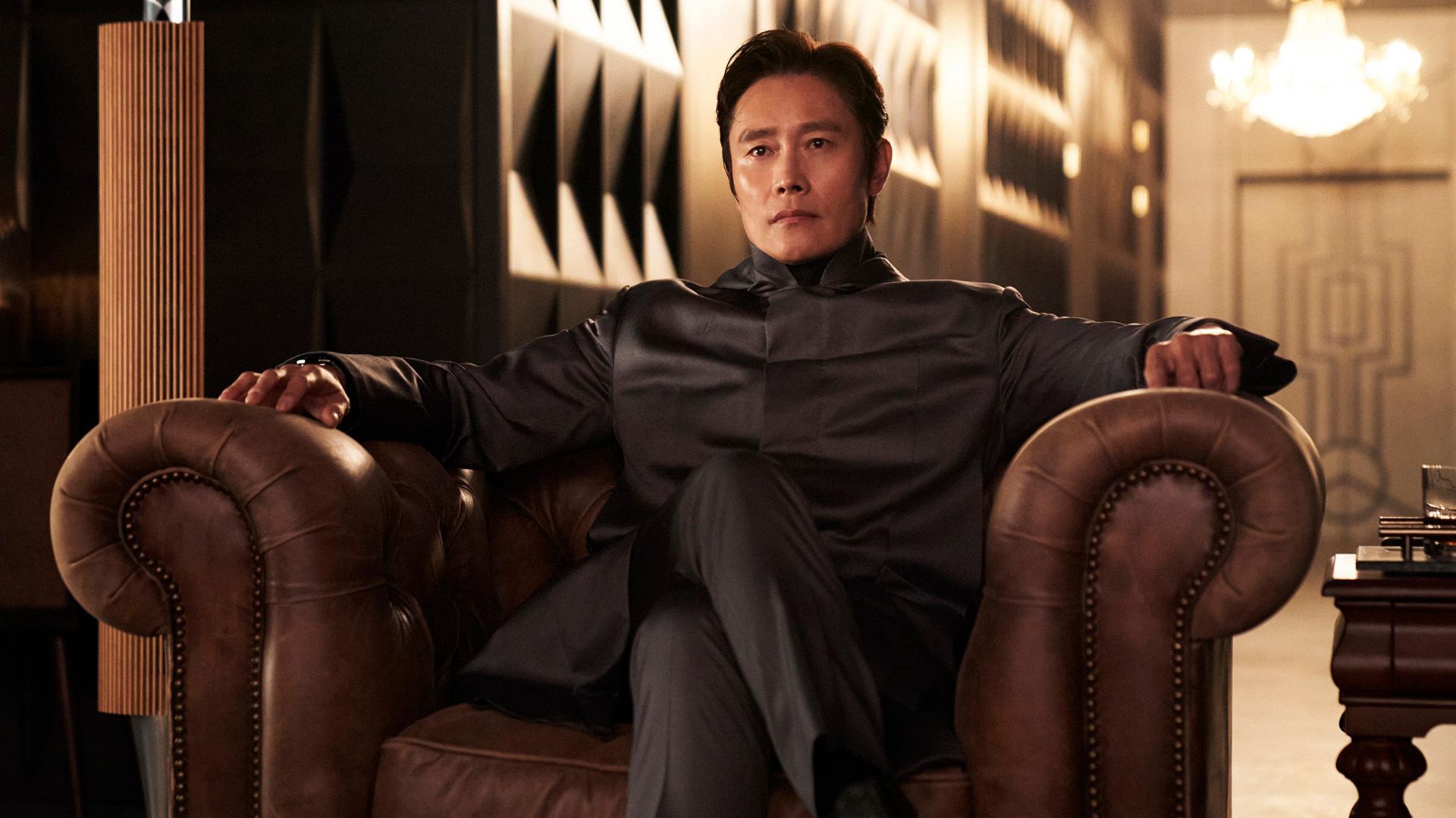 Lee Byung-hun in giacca grigia seduto su una poltrona di pelle con le gambe accavallate