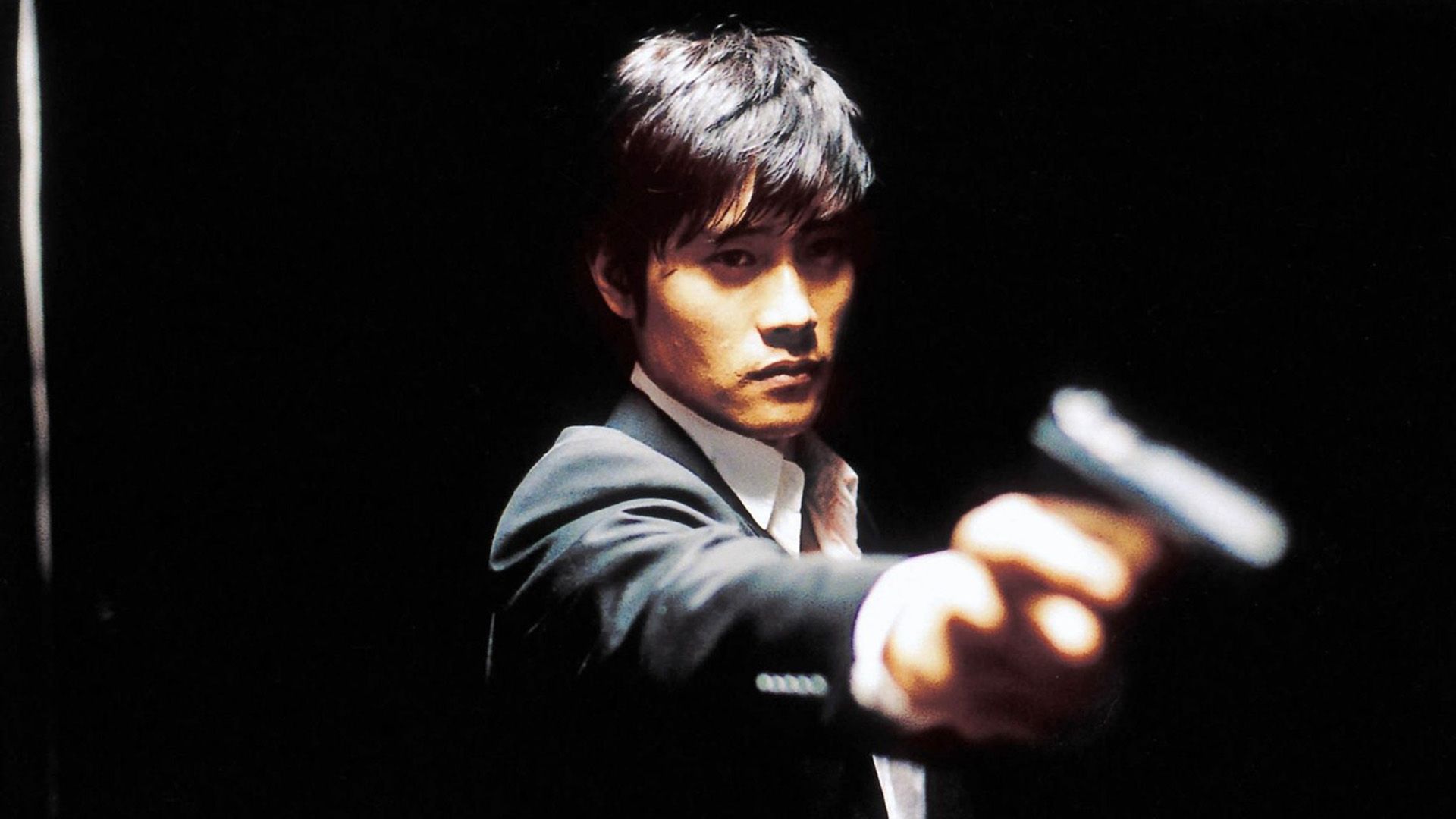 Lee Byung-hun in camicia bianca e giacca tiene una pistola con il braccio teso