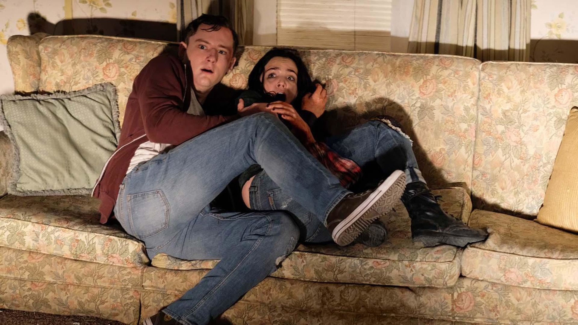 Lewis Pullman e una ragazza seduti terrorizzati sul divano con le gambe rannicchiate