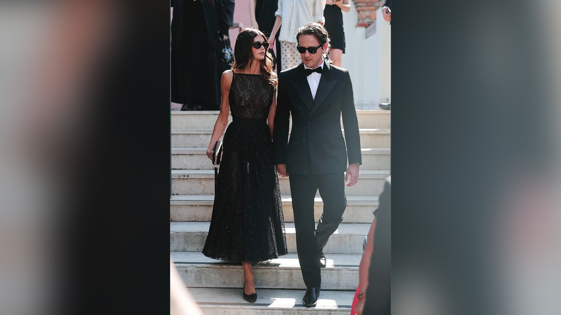 Lewis Pullman in smoking nero e Kaia Gerber in abito da sera nero scendono le scale, entrambi con occhiali da sole neri