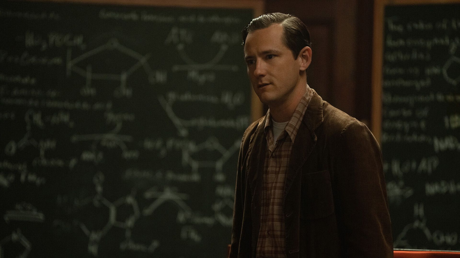 Lewis Pullman in blazer marrone di velluto a coste e camicia a quadri davanti a una lavagna con formule chimiche