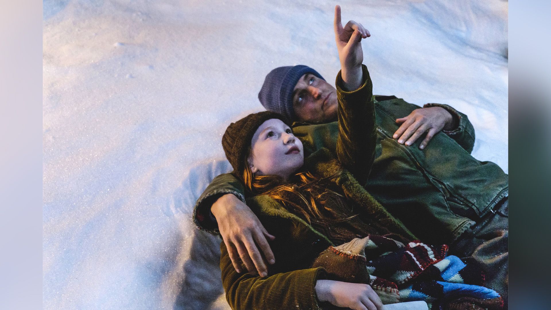 Ella Anderson e Woody Harrelson sdraiati sulla neve con Ella che indica il cielo