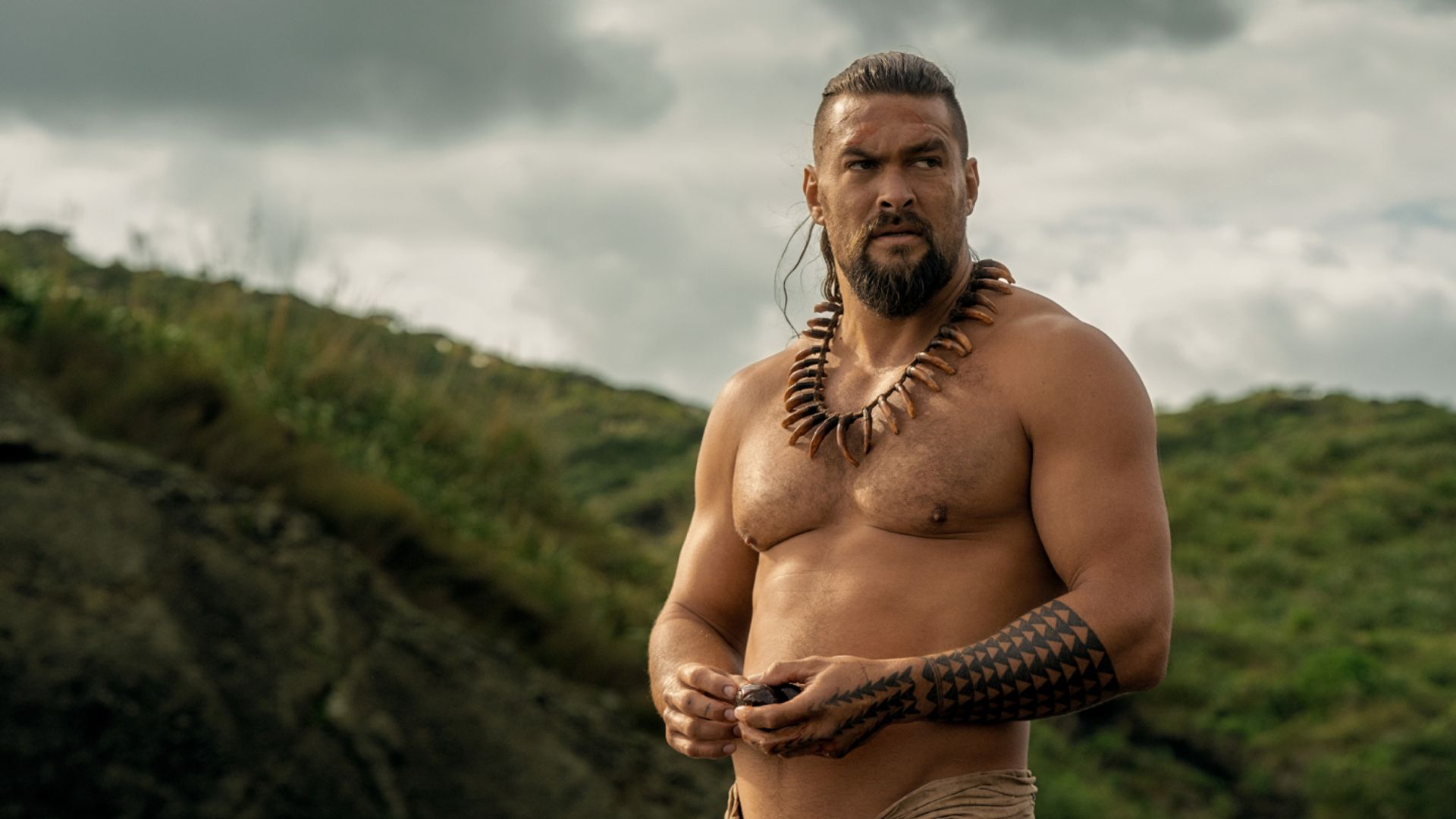 Jason Momoa a torso nudo con decorazioni tradizionali hawaiane e tatuaggi su sfondo montano