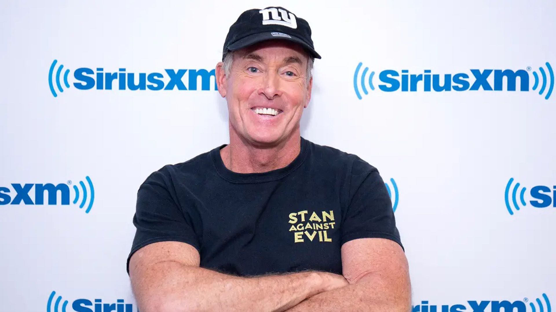 L'attore John C. McGinley con un berretto da baseball, in piedi a braccia conserte e con un ampio sorriso