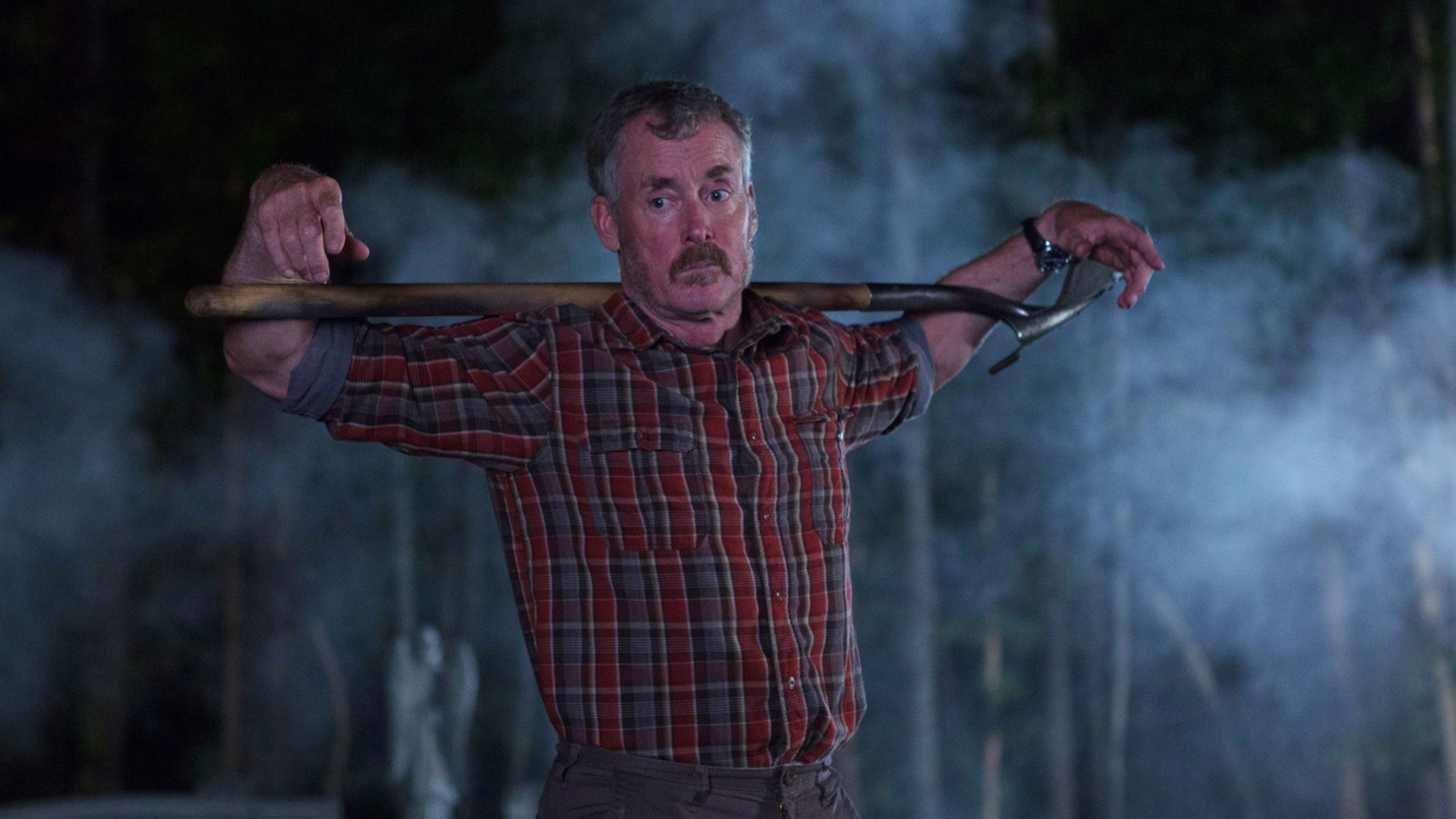 John C. McGinley in camicia a quadri rossa sta in piedi nella penombra con una pala sulle spalle